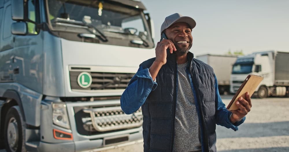 Smiling truck driver using a tablet at a freight yard, promoting transparent logistics services.