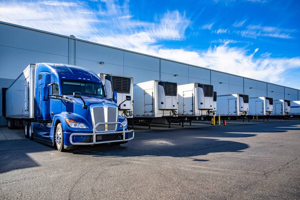 Blue semi-truck with multiple refrigerated trailers at a logistics hub.